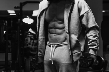 Fototapeta na wymiar A bodybuilder in his opened zipped hoodie is showing his abdominal muscles while holding dumbbells. A photo of a torso of a sporty guy who is posing after a workout in a gym.