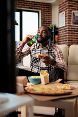 Cheerful man drinking alcoholic beer from bottle and eating takeaway meal from fast food delivery package, watching movie on tv. Enjoying drink and takeout meal in front of television.