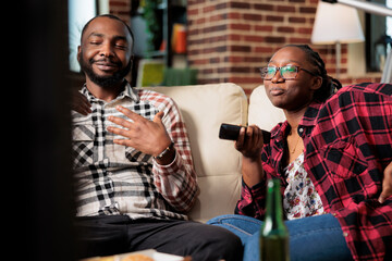 Life partners enjoying fast food delivery and watching tv, switching channels to find movie on television. Eating takeaway meal package and drinking beer from bottle, leisure activity.