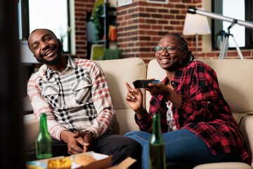 Cheerful couple laughing and watching television together, eating fast food delivery meal while girlfriend switches channels with tv remote control. Takeaway food and beer to have fun with film.