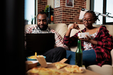 Man browsing internet on laptop and woman eating takeout food in front of television movie at home. Watching film on tv and using computer, eat takeaway dinner meal with bottles of beer.