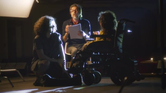 Male director of inclusive theater giving script recommendations to actress with a disability in wheelchair and actor during play rehearsal on a dim stage