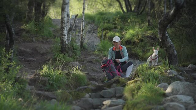 A female hiker sits on the rocky trail with her dog resting, checking her backpack. Slow-motion.