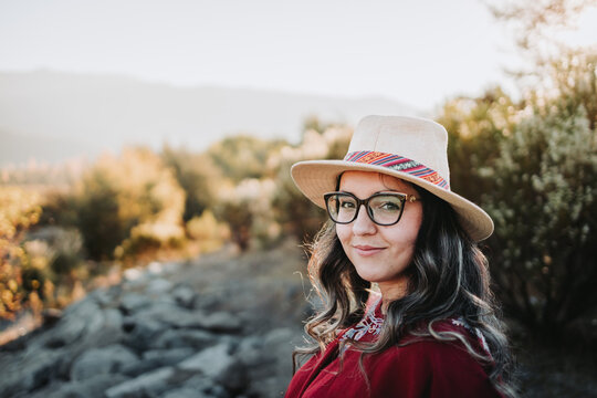Smiling latin backpacker woman wearing a traditional red poncho, glasses and a hat in a natural space at sunset
