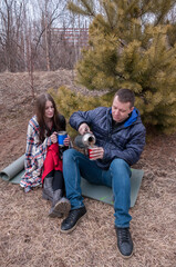 A couple in a spring park: a man and a girl sit near a fir tree and drink hot tea from a thermos