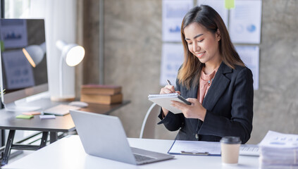 Young business Asian woman sitting at table and taking notes On table is laptop, smartphone and cup of coffee.On computer screen graphics and charts. 