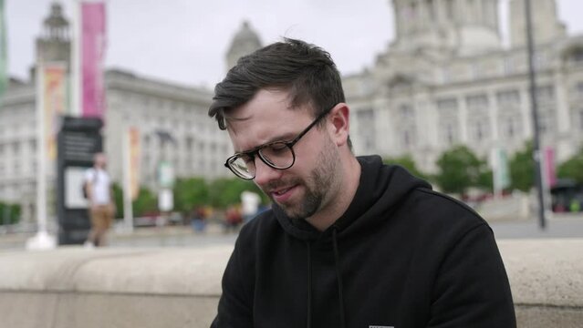 Young mid-twenties white male smiling using mobile phone answering call in left hand in cinematic slow motion. Mid-tight shot showing Liverpool in background.