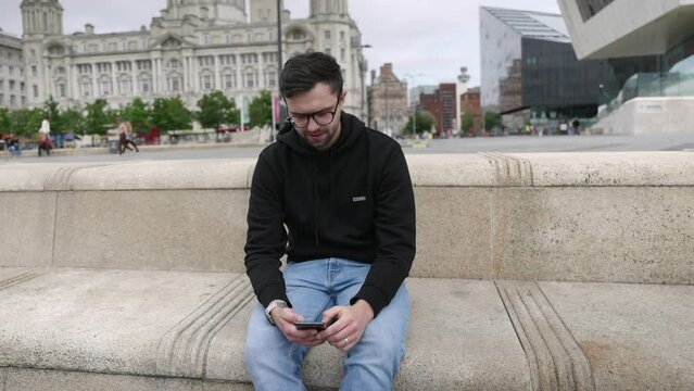Young mid-twenties white male using mobile phone in cinematic slow motion. Mid-wide shot showing Liverpool in background.