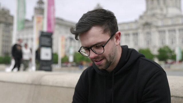 Young mid-twenties white male using mobile phone taking call in right hand in cinematic slow motion. Mid-tight shot showing Liverpool in background.