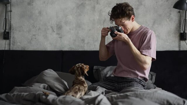 Young man is taking photo of dog and sitting on bed at home bedroom spbd. 4k American handsome guy holds camera and looks with smile, takes picture of cute pet and sits on blanket in room. One owner
