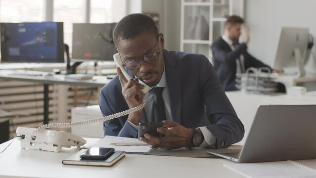 Slowmo of busy African American male broker in blue suit talking with investor on phone while monitoring online stock market on smartphone and laptop, sitting at desk in office with colleague