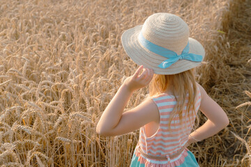 Child blonde girl in straw hat and nice dress smiling at the camera in wheat field
