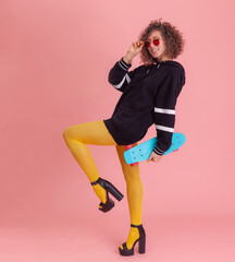 Stylish, bright, emotional cool style young woman dressed in yellow tights, sweater. Fashion Girl with curly hair and colored make-up posing with penny board on pink background