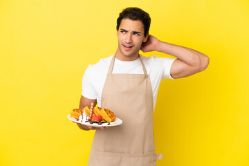 Restaurant waiter holding waffles over isolated yellow background having doubts