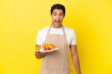 Restaurant waiter holding waffles over isolated yellow background with surprise facial expression