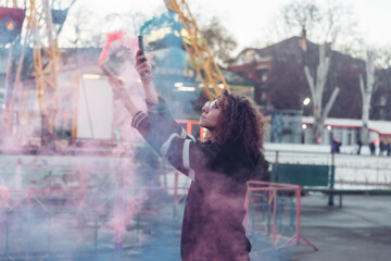 Bright cool woman with colored makeup and curly hair posing with colored smoke in the amusement park. Street fashion