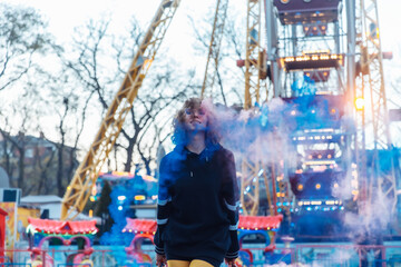 Bright cool woman with colored makeup and curly hair posing with colored smoke in the amusement park. Street fashion