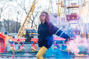 Fototapeta na wymiar Bright cool woman with colored makeup and curly hair posing with colored smoke in the amusement park. Street fashion