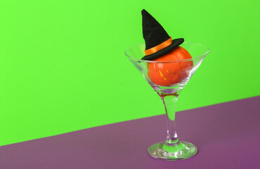 Halloween party. Halloween pumpkin with witch hat in cocktail martini glass on green purple background. Minimal layout