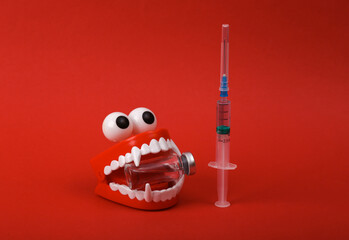 Funny toy clockwork jumping teeth with eyes and vaccine bottle, syringe on red background. Vaccination