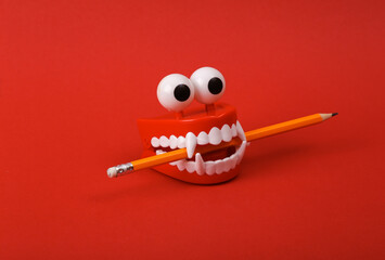 Funny toy clockwork jumping teeth with eyes holding pencil on red background.