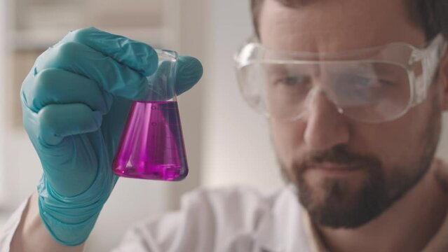 Slowmo closeup of Caucasian male chemist in protective goggles and lab coat looking at purple liquid in glass flask