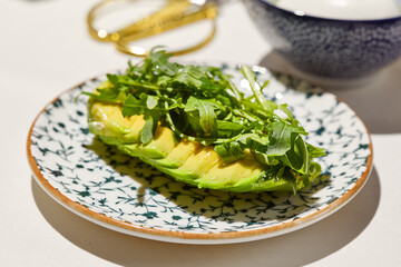 Veggie appetizer from avocado and arugula on ceramic plate on white table. Avocado starter with rocket salad in vegan menu. Appetizer from sliced avocado and greens. Healthy food. Vegetarian dish.