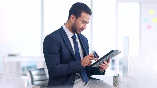 Male executive online shopping, buying with a corporate credit card. A smiling man paying business bills, sitting on an office desk. Happy guy making a purchase on a digital tab.