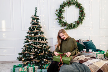 A young woman opens Christmas gifts in a New Year's atmosphere and rejoices. The woman is in a bright, cozy bedroom, beautifully decorated for Christmas.