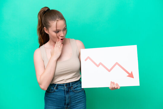 Young caucasian woman isolated on green background holding a sign with a decreasing statistics arrow symbol with surprised expression
