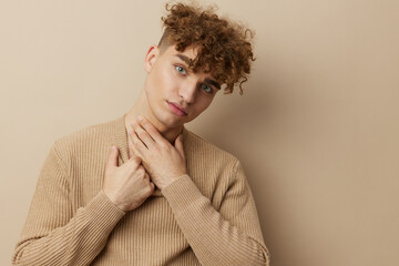 a charming young man with curly hair stands on a beige background in a stylish sweater and looks at the camera with a slight smile, relaxed touching his neck with his hand
