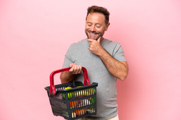 Middle age caucasian man holding a shopping basket isolated on pink background looking to the side...