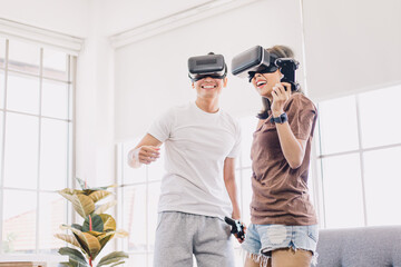 Young Asian couple playing and enjoying video games with VR head sets at home. Happy couple having fun with virtual reality glasses in living room. 