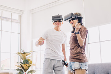 Young Asian couple playing and enjoying video games with VR head sets at home. Happy couple having...