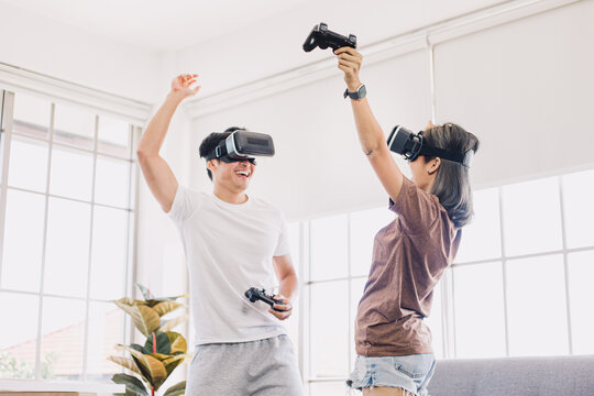 Young Asian couple playing and enjoying video games with VR head sets at home. Happy couple having fun with virtual reality glasses in living room.
