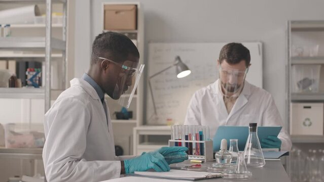Medium slowmo of two diverse male chemists wearing lab coats and protective face shields sitting at table with glass equipment on it and doing research with substances in modern laboratory