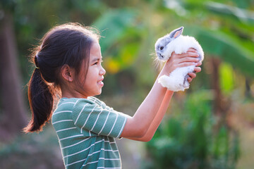 Asian child girl holding and hugging her adorable bunny fluffy with tenderness and love. Kid take care and play with pet in the garden. Symbol of Easter day.