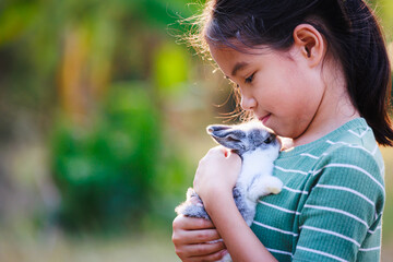 Asian child girl holding and hugging her adorable bunny fluffy with tenderness and love. Kid take care and play with pet in the garden. Symbol of Easter day.