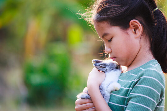 Asian child girl holding and hugging her adorable bunny fluffy with tenderness and love. Kid take care and play with pet in the garden. Symbol of Easter day.