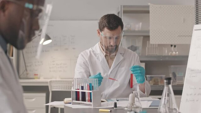 Medium slowmo of Caucasian male chemist in workwear dropping red liquid substance from test tube to petri dish with pipette doing research, working in scientific lab with African American colleague