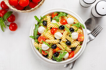 Italian cold pasta salad. Pasta fredda alla caprese. Fusilli, tomato, mozzarella, olive, arugula. Directly above on a white table surface.