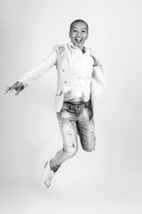 Fototapeta premium Beauty, style, business and fashion concept. Studio portrait of happy Thai woman with bright clothes, suit and torn jeans jumping in air. Model with shaved head. Black and white image with copy space