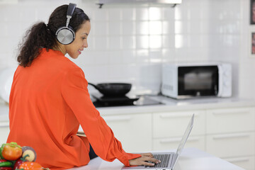 Happy latin woman learn cooking food while checking recipe from internet with computer in kitchen. Young female with headphone prepare delicious meal and listen music. Healthy and lifestyle concept