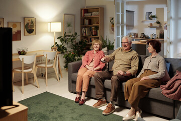 Three aged friends in casualwear sitting on sofa in front of tv set while man with remote control in hand choosing channel