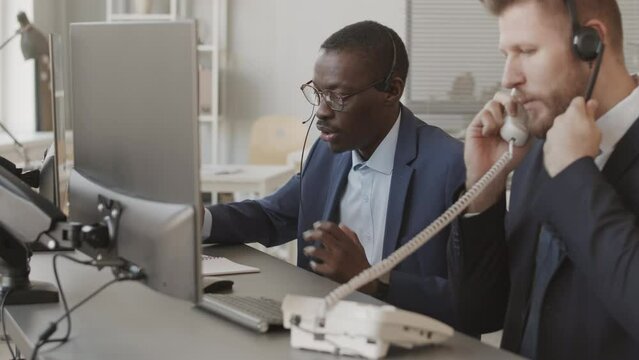 Two ambitious multiethnic stock brokers wearing suits and headsets following online stock market on their computer monitors while talking to clients