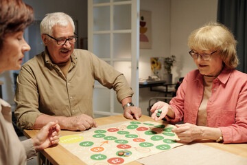 Happy mature woman throwing dice cubes over paper board with green and red circles of leisure game...