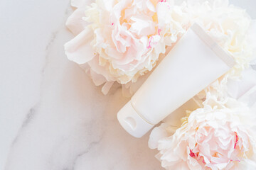 Flat lay composition with cosmetic products and peony flowers on a marble. Top view. Unbranded plastic white tube with face or hand cream or cosmetic mask