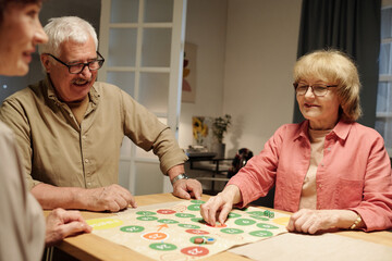 Group of happy aged friends playing board game while one of them moving chip from one circle to...