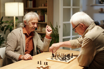 Happy senior man looking at his buddy moving black figure along chessboard while both playing chess by table at leisure