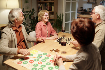 Cheerful aged woman laughing during name game with friends gathered by table with chessboard and paper with green and red circles